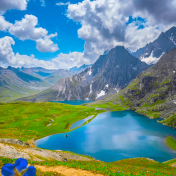 A breathtaking view of the serene Vishansar Lake, surrounded by lush green meadows and snow-capped peaks, captured during the Kashmir Great Lakes Trek. The clear blue sky reflects on the pristine waters, creating a perfect mirror image of the majestic mountains.
