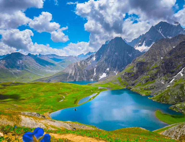 A breathtaking view of the serene Vishansar Lake, surrounded by lush green meadows and snow-capped peaks, captured during the Kashmir Great Lakes Trek. The clear blue sky reflects on the pristine waters, creating a perfect mirror image of the majestic mountains.
